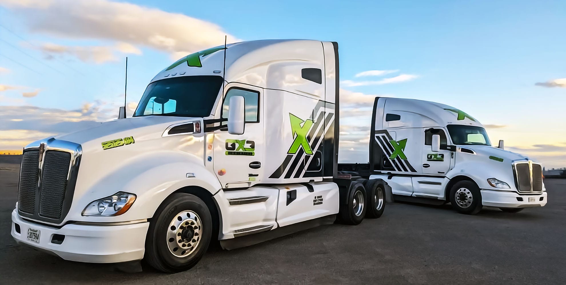 Two white semi-trucks with green X graphics parked side by side under a clear sky